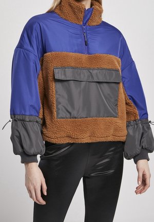 Femme portant une veste color-block avec des manches bleues, un corps en polaire marron, une grande poche avant grise et des leggings noirs brillants.