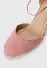 Chaussure à talon haut en daim rose avec un bout pointu et des côtés ouverts. Présente une fine lanière à la cheville et une semelle intérieure lisse beige.