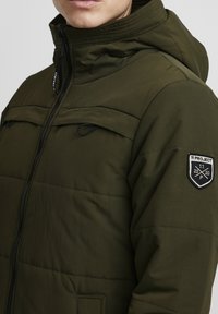 Hooded, olive green puffer jacket with a zip closure, two chest pockets, and a round patch on the left sleeve featuring a logo.