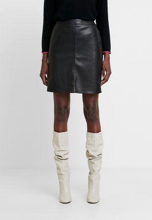 Mujer con minifalda de cuero negra, top de manga larga negra y botas blancas desgarbadas hasta la rodilla sobre un fondo liso.