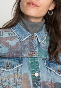 Kvinna som bär silver hoops, en grå polotröja och en patchwork-jeanjacka med tryckta mönster och metallknappar.