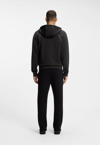 Mann steht mit dem Rücken zur Kamera, trägt einen schwarzen Kapuzenpullover mit reflektierenden Paspeln und schwarze Hose vor einem schlichten weißen Hintergrund.