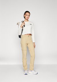 Pantaloni cargo beige con tasche con zip, abbinati a una maglietta bianca a maniche lunghe e corte con un piccolo logo. Sneakers bianche con dettagli colorati.