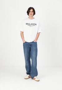 Balta pārāk liela T-kreklu ar pelēku tekstu "McLaren Formula 1 Team", saskaņota ar ziliem džinsiem un baltiem apaviem ar sarkaniem svītrojumiem.