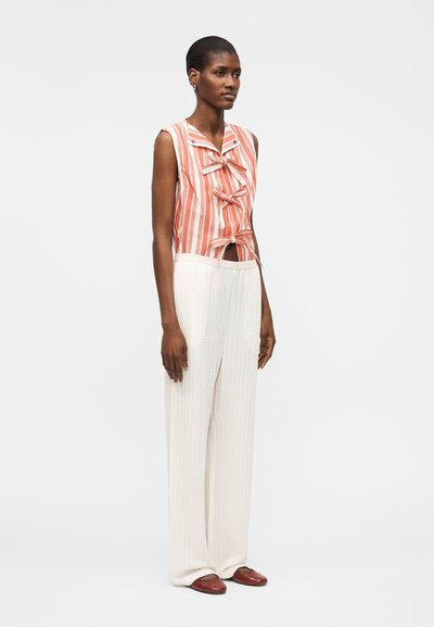 Γυναίκα στέκεται φορώντας ένα πορτοκαλί και λευκό ριγωτό αμάνικο τοπ με φιόγκους και φαρδιά λευκά υφασμάτινα παντελόνια με καφέ παπούτσια.