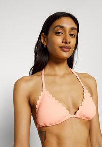 Top de biquíni triangular em rosa coral, com bordas rendadas, laços halter e um detalhe franzido na frente para um visual texturizado.