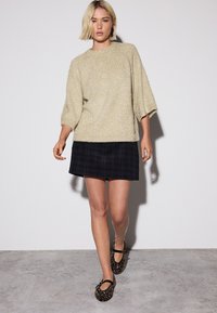 Cremefarbener, strukturierter Pullover mit 3/4-Ärmeln, kombiniert mit einem schwarzen karrierten Minirock. Schwarze gemusterte Ballerinas vervollständigen das Outfit.