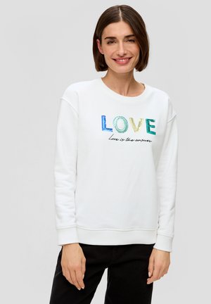 Donna con capelli corti castani che sorride, indossa una felpa bianca con lettere multicolori "LOVE" e il testo "l'amore è la risposta", pantaloni neri.