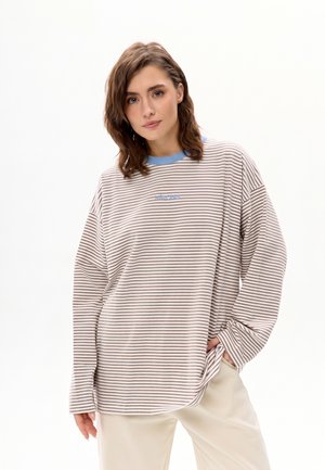 Jeune femme aux cheveux bruns mi-longs, portant une chemise ample beige rayée à manches longues avec un col bleu et un pantalon clair, debout avec une main dans la poche.