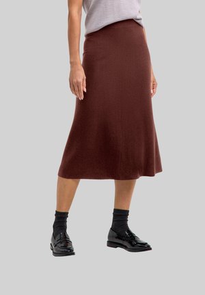 Femme portant une jupe midi marron associée à des chaussettes noires et des mocassins noirs brillants, debout devant un fond gris clair uni.