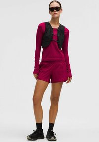 Aansluitend magenta shirt met lange mouwen en duimopeningen, gecombineerd met magenta shorts. Laagjes zwarte lichtgewicht vest met sluitingen, zwarte sportschoenen.