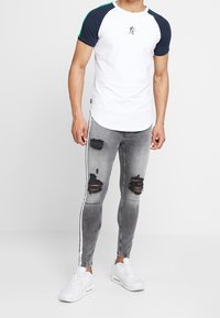 Vit t-shirt med marinblå ärmar, rund halsringning och logotyp. Grå slitna jeans med hål och vita sidostråk. Vita sneakers.