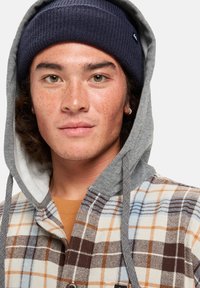 Camisa de franela multicolor con capucha gris, que presenta un estampado a cuadros en marrón, beige y azul, con un gorro de lana azul oscuro y botones visibles.