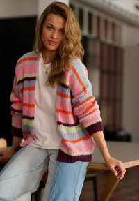 Mladá žena s vlnitými vlasy sedí na okraji stolu, na sobě má barevný pruhovaný kardigan, bílou košili a světle modré džíny uvnitř.