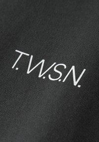 Fekete szövet, amelyen fehér betűzés található, ami a "T.W.S.N." feliratot tartalmazza, texturált felülettel és letisztult, modern betűstílussal.