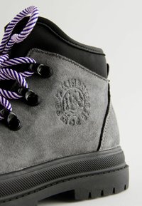 Bottine en daim gris avec des accents noirs, lacets rayés violets et blancs, détail de logo en relief, et semelle en caoutchouc noir texturé.