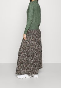 Un pull en tricot vert avec un motif texturé, associé à une jupe maxi noire à fleurs avec de petites fleurs roses et blanches, et des baskets blanches.