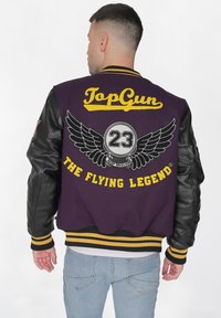 Violetti college-takki mustilla nahkahihnoilla, jossa on keltainen "TopGun" -teksti, brodeerattu siipilogo ja harmaa "23", raidalliset hihansuut.