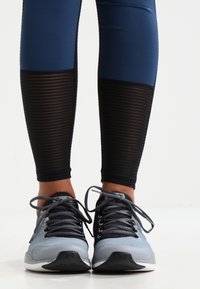 Leggings blu con accenti a strisce nere, abbinati a scarpe da ginnastica grigie con rete grigia e lacci scuri. Sfondo con superficie piatta.