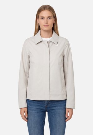 Donna con una giacca beige chiaro con colletto, abbinata a jeans blu, in piedi davanti a uno sfondo bianco uniforme.