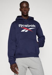 Hanorac Reebok de culoare bleumarin, fabricat din bumbac, având un logo mare alb cu accente roșii, glugă cu șnur și buzunar tip kangaroo în față.