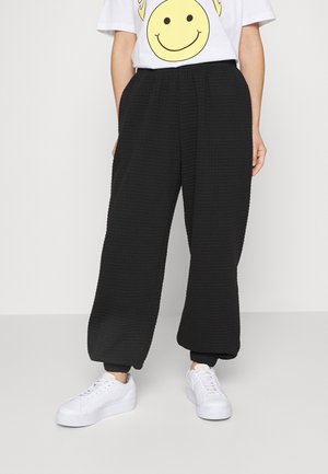 Person trägt schwarze strukturierte Hose mit weitem Bein, weiße Sneakers und ein weißes T-Shirt mit großem gelbem Smiley-Gesicht.