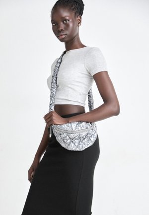 Nuori nainen, jolla on letitetyt hiukset, pukeutuu harmaaseen lyhyeen toppiin ja mustiin hameeseen, pitäen hopeista tiiliskuvioista olkalaukkua valkoista taustaa vasten.