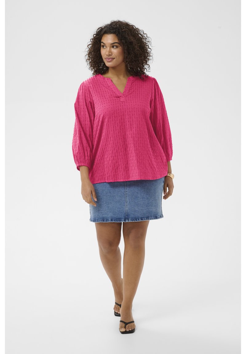 Femme aux cheveux bouclés portant un chemisier rose texturé, une jupe en denim bleu et des sandales noires, debout avec un léger sourire et regardant sur le côté.