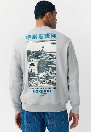 Šedá mikina s grafickým motívom na zadnej strane, z čierno-bieleho japonského umenia, modrý nápis „Hokusai“ a „Kajikazawa v provincii Kai.“