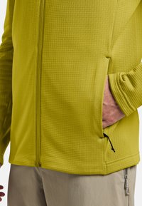 Veste zippée vert lime en tissu texturé, avec poches latérales et ourlet ajustable. Elle possède des manches longues et une finition lisse.