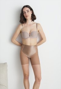 Sujetador beige sin costuras con aros y tirantes ajustables, combinado con pantalones cortos modeladores marrones de cintura alta y transparentes. Material suave y ligero.