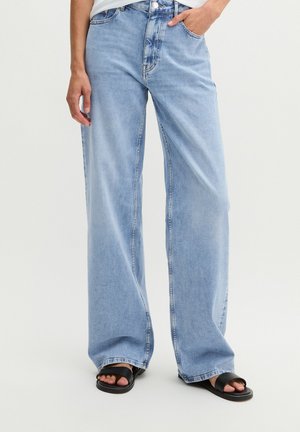 Person trägt hellblaue Jeans mit weitem Bein, eine Hand in der Tasche und schwarze offene Sandalen, steht vor einem einfachen hellen Hintergrund.