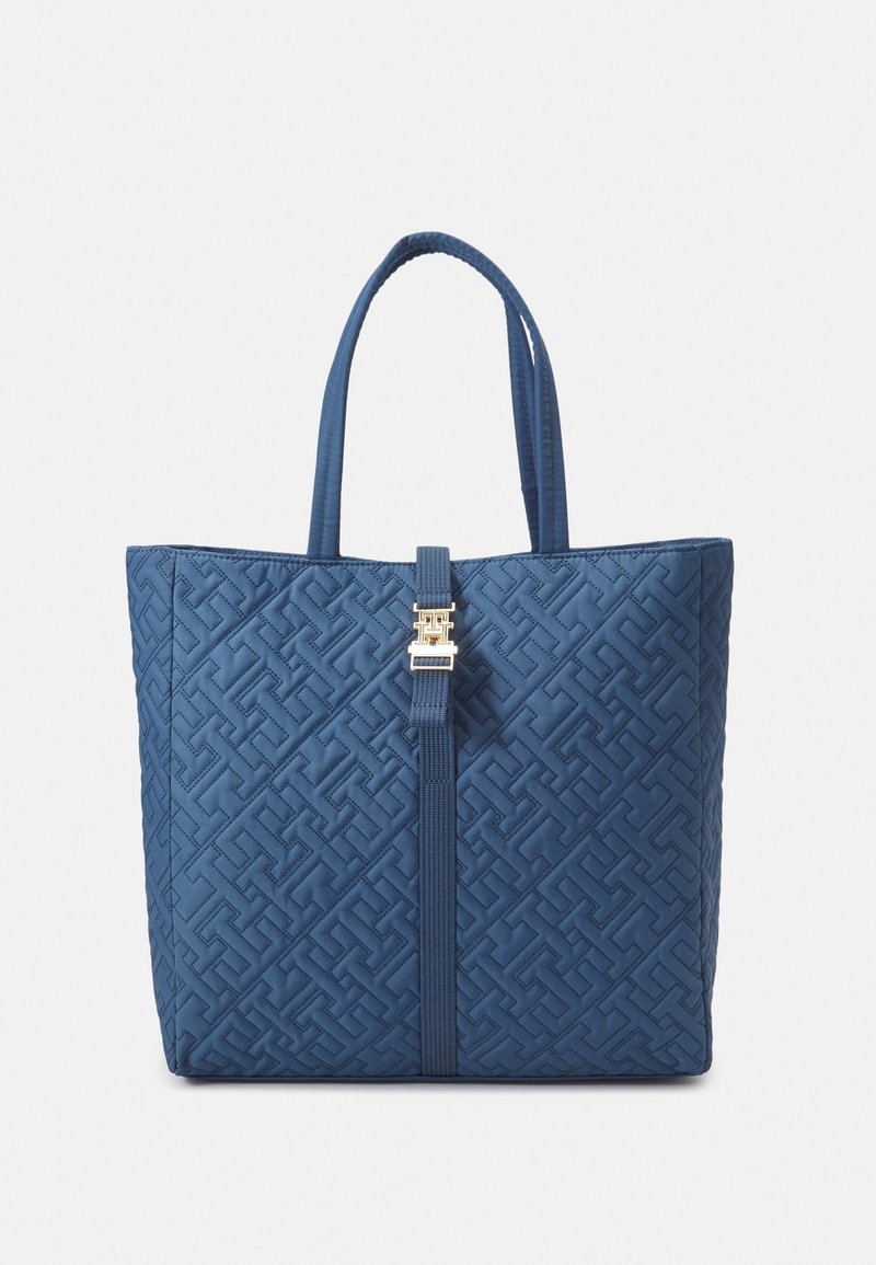 Sac cabas matelassé bleu avec doubles poignées et une sangle centrale ornée d'une boucle décorative dorée sur un fond uni.