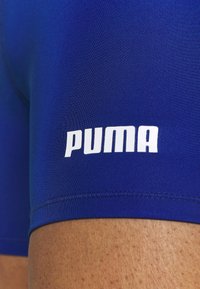 Gros plan d’un vêtement de sport bleu montrant le logo blanc Puma sur le tissu au-dessus de la cuisse d’une personne.