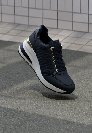 Navy blue athletic shoe with quilted texture, gold eyelets and accents, white midsole, and a cushioned collar. Suitable for casual wear.