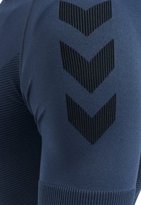 Närbild av blå tygbyxor i leggingsstil med tre svarta chevronränder på låret och ribbat mönster nära knäet.