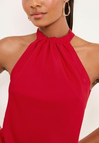 Sarkans haltera tops ar koptu apkakli, izgatavots no mīksta, gluda auduma. Tajā ir minimālistisks dizains un tas atklāj plecus.