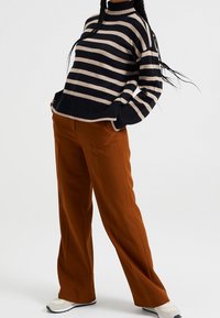 Černý a béžový pruhovaný svetr s vysokým výstřihem, žebrovanými manžetami a volným střihem, v kombinaci s hnědými širokými kalhotami a bílými teniskami.