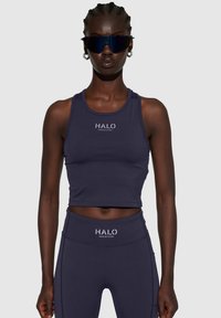 Marineblauwe cropped tanktop met HALO-logo; glad, rekbaar materiaal; gecombineerd met bijpassende high-waisted leggings. Donkere zonnebril als accessoire.