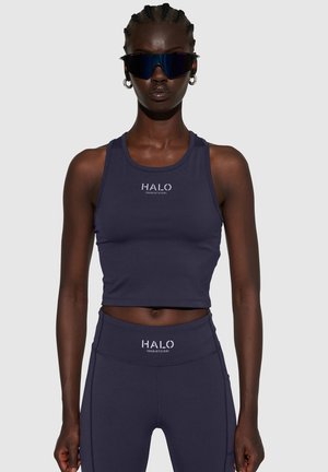 Marineblauwe cropped tanktop met HALO-logo; glad, rekbaar materiaal; gecombineerd met bijpassende high-waisted leggings. Donkere zonnebril als accessoire.