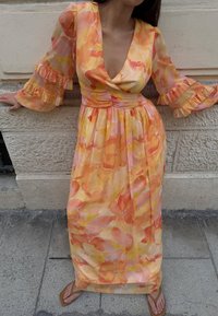 Eine Frau trägt ein langes, fließendes Kleid mit aquarellartigen Mustern in Orange, Gelb und Pink, einem tiefen V-Ausschnitt und rüschierten Glockenärmel, während sie vor einer Steinmauer steht.
