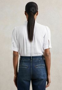 Weißes kurzärmliges Bluse mit Falten und Knopfakzenten, kombiniert mit hoch taillierten dunkelblauen Jeans aus Denim mit zwei Gesäßtaschen.