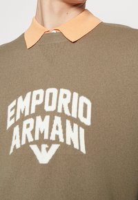 Pull en tricot vert olive avec logo blanc "EMPORIO ARMANI", présentant une finition texturée et un col côtelé orange.