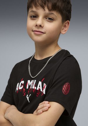 Czarna bawełniana koszulka z napisem „AC Milan” w kolorze białym i czerwonym z płomieniami, krótkimi rękawami oraz czerwoną naszywką z logo na lewym rękawie. Srebrny łańcuszek na szyi.