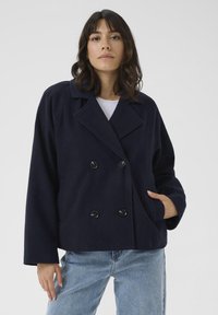 Tamsiai mėlynas dvigubo užsegimo vilnonis švarkas su plačiu apykakle, dideliais juodais mygtukais ir šoninėmis kišenėmis. Atsipalaidavęs siluetas ir minkšta tekstūra.