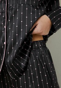 Zwart pyjamapakket in satijnen stof met dunne roze hartpatronen en verticale lijnen. Bevat een blouse met kraag en een broek met elastische tailleband.