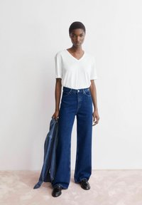 Weißes V-Ausschnitt-T-Shirt, weiter dunkelblauer Jeans mit weitem Bein, hält eine Jeansjacke, kombiniert mit schwarzen Schuhen, vor einem hellen Hintergrund.