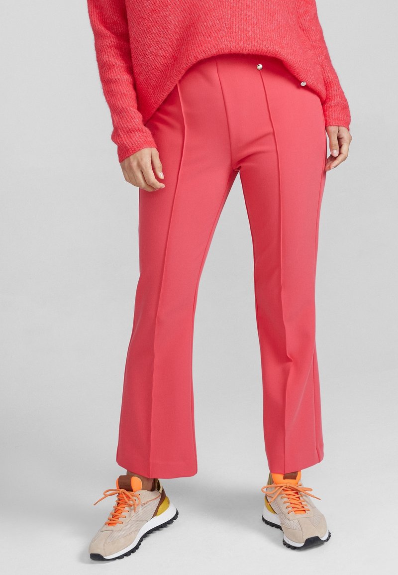 Pantalons corail ajustés avec une finition texturée, dotés de plis à l'avant et de boutons décoratifs. Associés à des baskets beiges et des lacets orange vif.