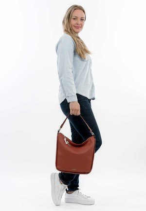 Brown leather shoulder bag with a smooth texture, rounded shape, and a detachable strap, held by a person in casual attire.