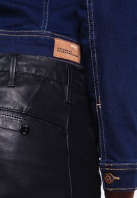 Svarta läderbyxor med framficka, kombinerat med en blå denimjacka med kontrastsömmar och en brun etikett med nitar.